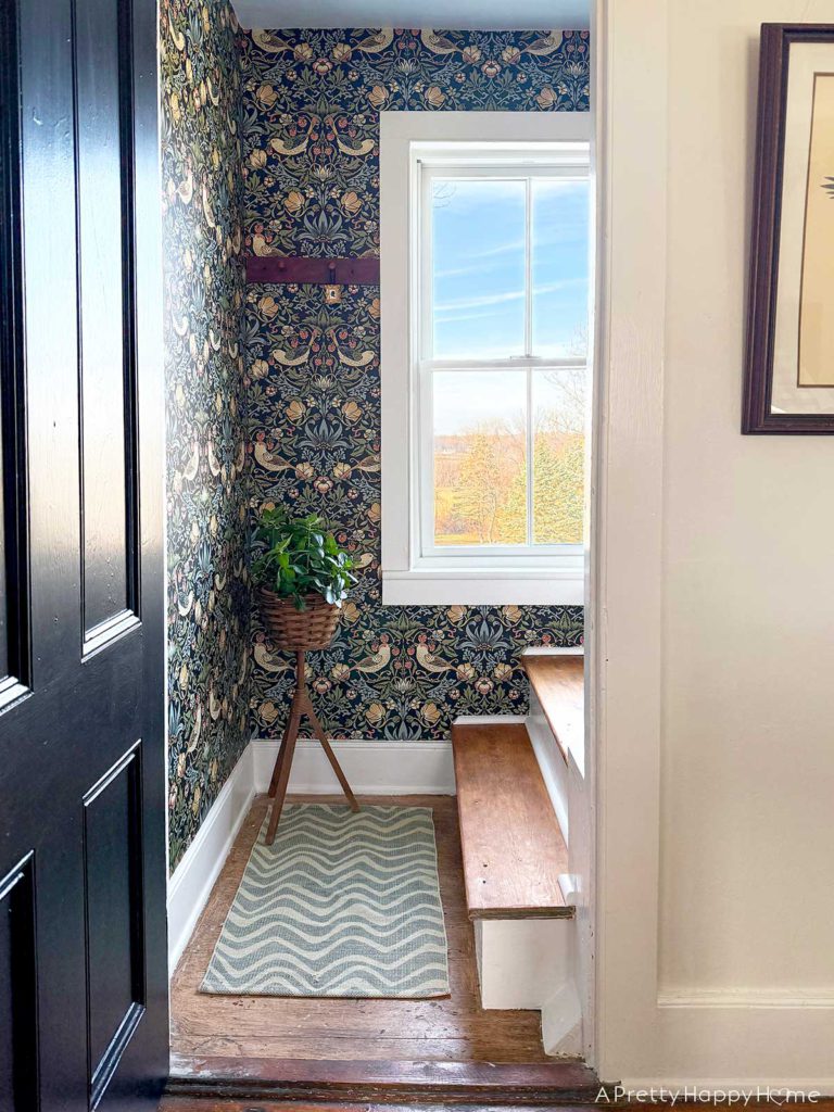 black door opening to colorful william-morris style wallpaper in a tiny stairwell with wood stairs. There a red peg rail on the wall and below the stairwell window is a basket with a plant in it
