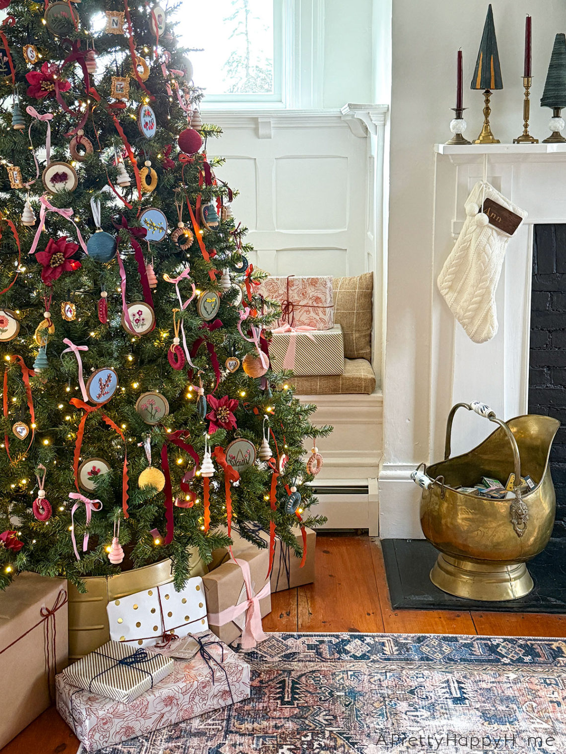 christmas tree decorated with colorful ribbons and hand embroidered flower ornaments set in front of a built in, white bench next to a fireplace with white mantel in a 250 year old home. On the fireplace are burgundy candles and small green christmas trees. Colorful packages are wrapped under the tree. Featured on the Happy List.
