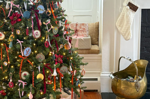 christmas tree decorated with colorful ribbons and hand embroidered flower ornaments set in front of a built in, white bench next to a fireplace with white mantel in a 250 year old home. On the fireplace are burgundy candles and small green christmas trees. Colorful packages are wrapped under the tree. Featured on the Happy List.