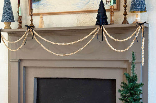 grey fireplace with a snowy christmas painting hanging over the mantel. there are natural wood bead garlands draped in in front of the mantel. Sittin got the mantel are a variety of mini christmas trees in a blue color, and a handful of burgundy candles in brass candles holders. Image featured on the happy list
