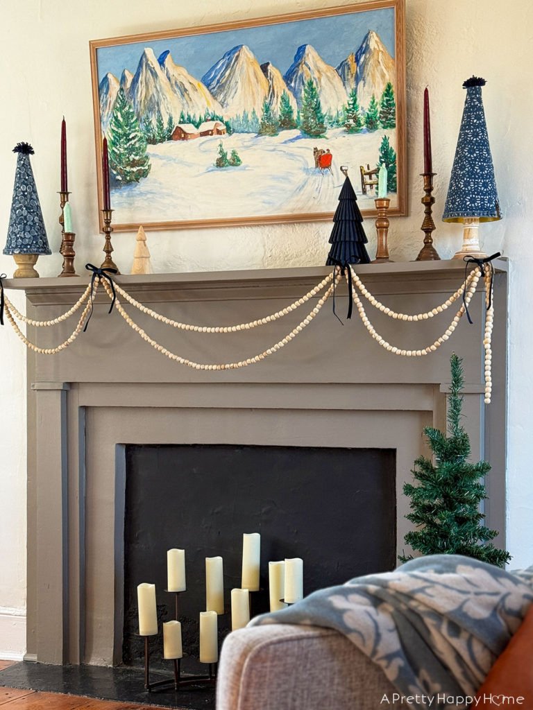 grey fireplace with a snowy christmas painting hanging over the mantel. there are natural wood bead garlands draped in in front of the mantel. Sittin got the mantel are a variety of mini christmas trees in a blue color, and a handful of burgundy candles in brass candles holders. Image featured on the happy list
