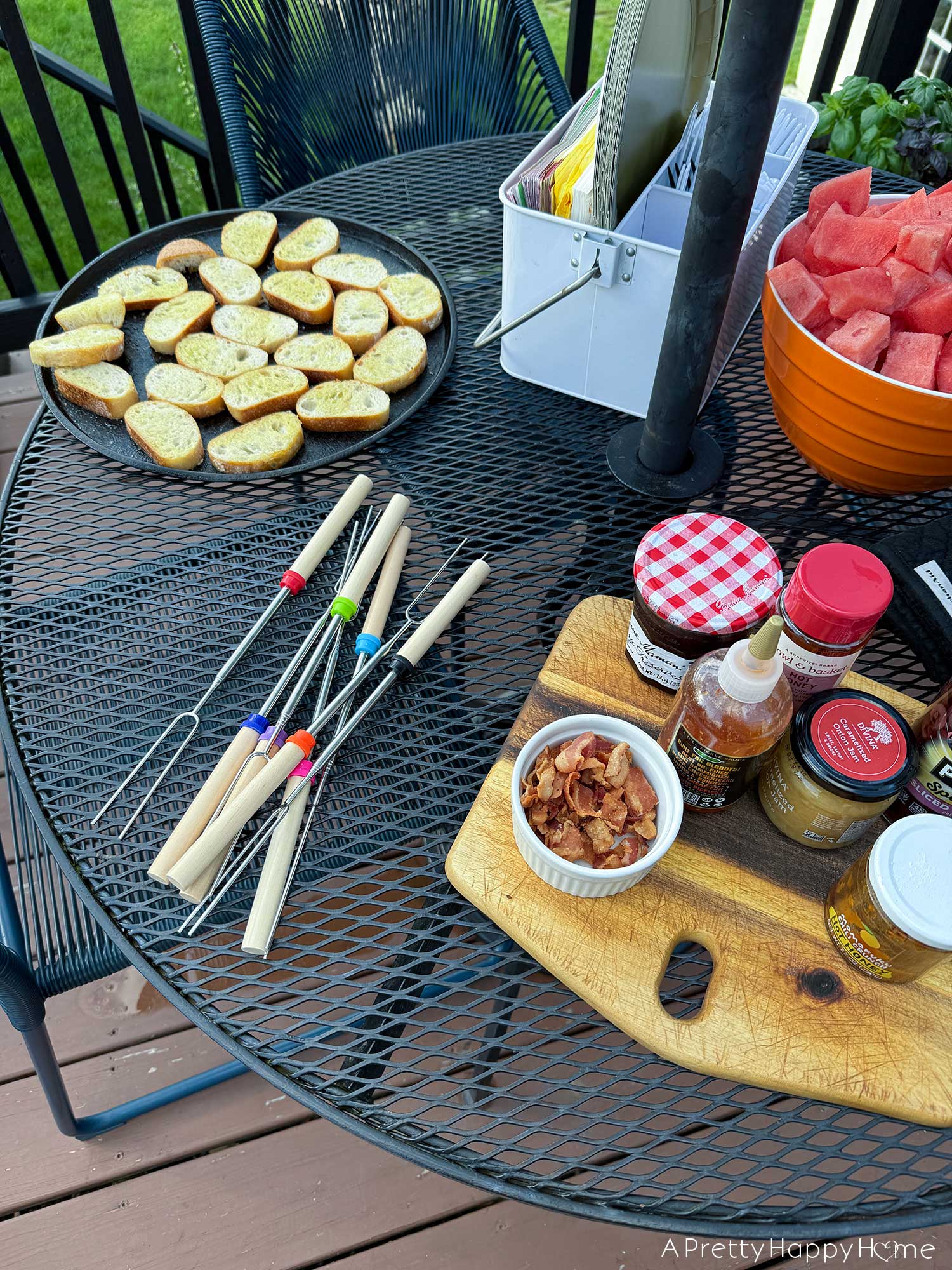 sliced bread, watermelon, and a board of condiments sits next to colorful roasting sticks on a black table
