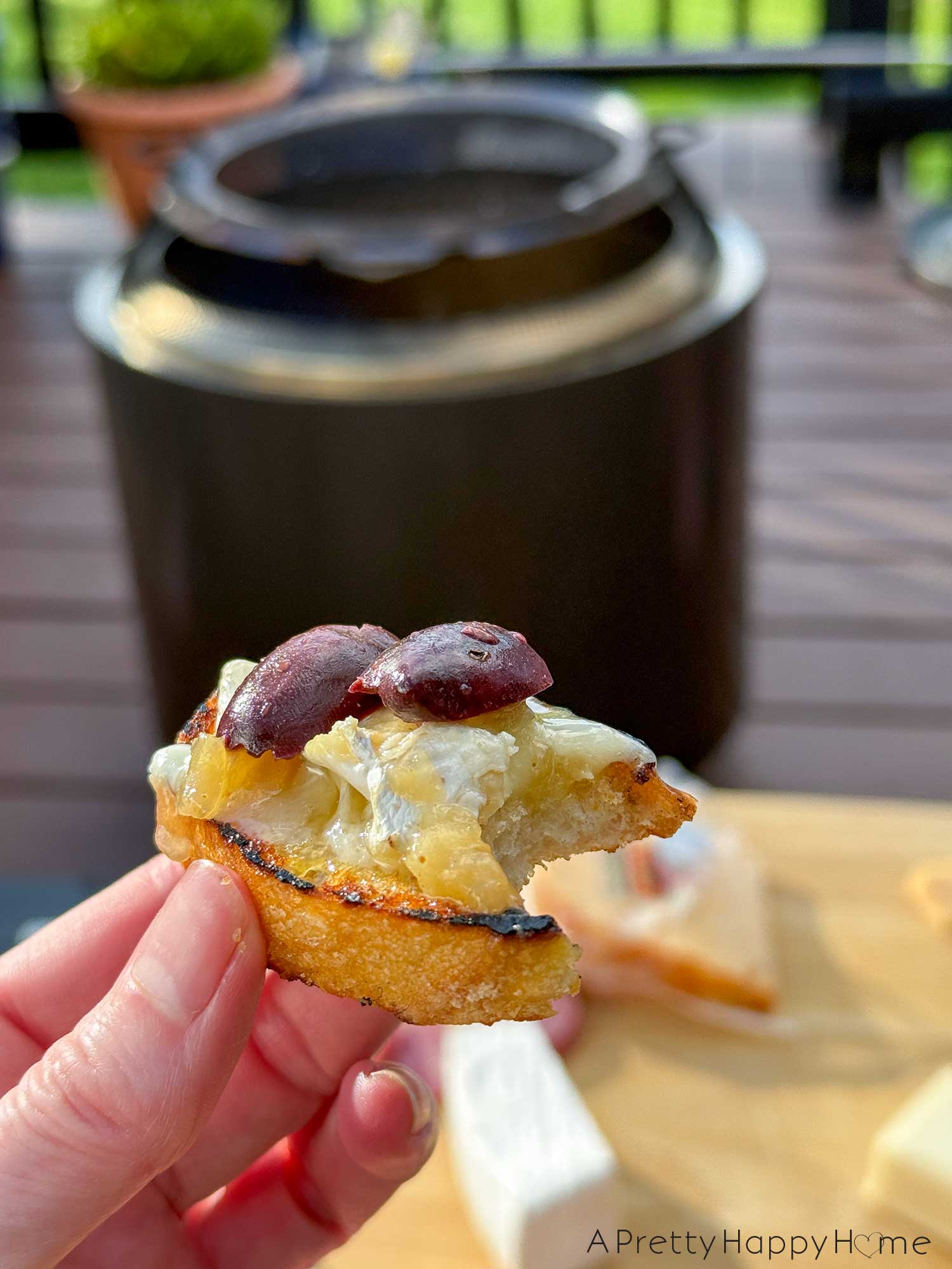 woman's hand holding a savory s'more made with crostini topped with melted cheese, onion jam, and kalamata olives. In the background is a solo stove fire pit in black.