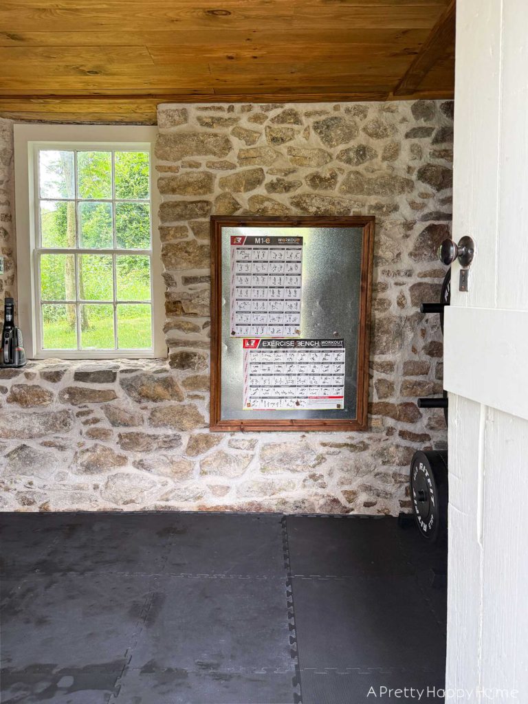 Home gym with fieldstone walls, wood ceiling, and black cushioned floor. Hanging on the wall to the right of a large single-paned old window is a bulletin board with posters showing weightlifting exercises.