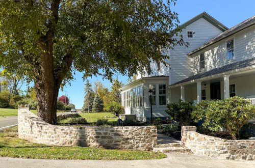 colonial farmhouse from the 1780s painted white standing behind a curved fieldstone wall