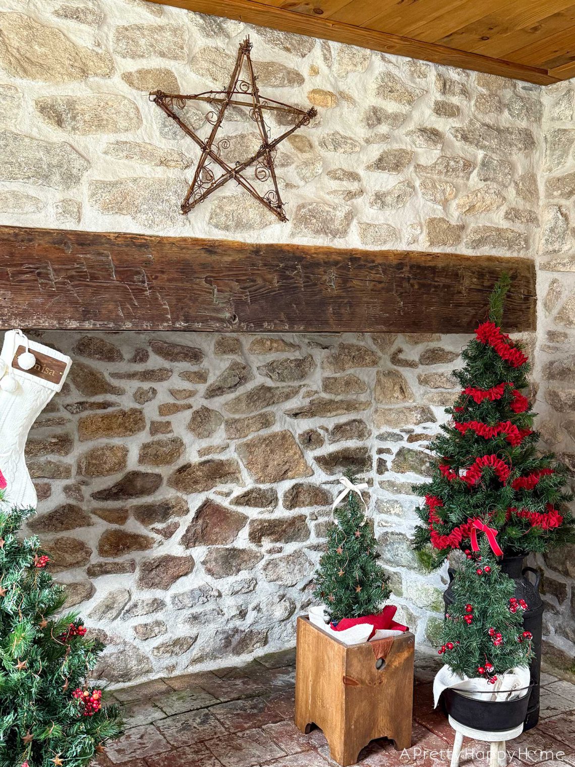 stone fireplace with wood mantel shown with a rustic twig star above the mantel. There are 3 trees decorated with red garland and ribbon in front of the fireplace. Shown on the happy list.