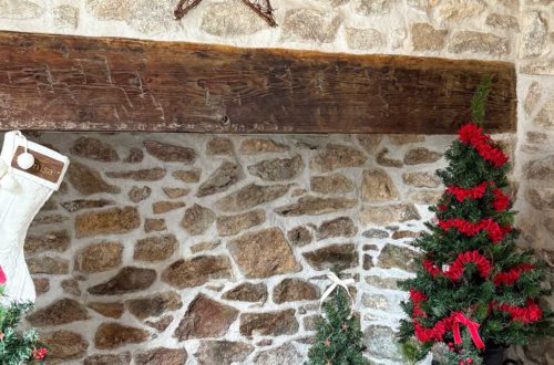 stone fireplace with wood mantel shown with a rustic twig star above the mantel. There are 3 trees decorated with red garland and ribbon in front of the fireplace. Shown on the happy list.