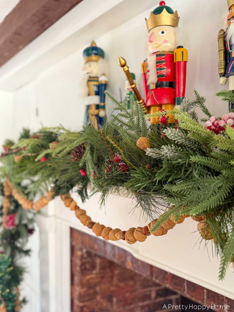 Large fireplace in an old house decorated for christmas with seven nutcrackers of varying sizes on the mantel and an off centered green garland with red berries. Layered on the mantel is a handmade nut garland comprised of walnuts, hazelnuts, and almonds. It is beautiful festive nutcracker Christmas mantel decor.