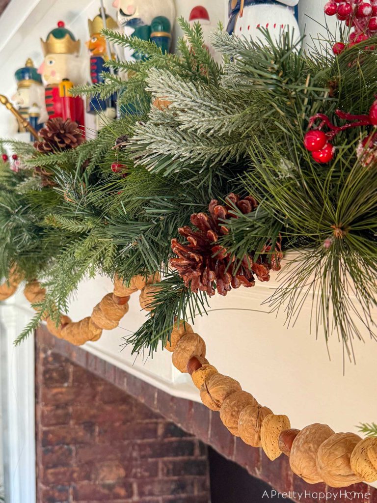 green holiday garland adorning a white fireplace mantel with brick surround. There is a hand strung, in-shell nut garland hanging below the green garland. On top of the mantel are colorful nutcrackers. All a part of the blog post five ways to decorate with nuts for christmas.
