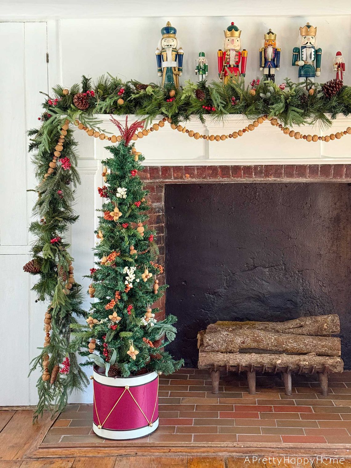 large fireplace in an old house decorated for christmas with seven nutcrackers of varying sizes on the mantel and an off centered green garland with red berries. Layered on the mantel is a handmade nut garland comprised of walnuts, hazelnuts, and almonds. Sitting in front of the fireplace is a skinny christmas tree with a tree collar that looks like a red drum. The tree is decorated with faux red and white berries and ornaments fashioned out of nuts.