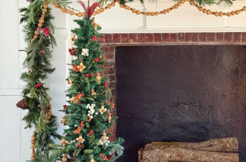 large fireplace in an old house decorated for christmas with seven nutcrackers of varying sizes on the mantel and an off centered green garland with red berries. Layered on the mantel is a handmade nut garland comprised of walnuts, hazelnuts, and almonds. Sitting in front of the fireplace is a skinny christmas tree with a tree collar that looks like a red drum. The tree is decorated with faux red and white berries and ornaments fashioned out of nuts.