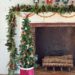 large fireplace in an old house decorated for christmas with seven nutcrackers of varying sizes on the mantel and an off centered green garland with red berries. Layered on the mantel is a handmade nut garland comprised of walnuts, hazelnuts, and almonds. Sitting in front of the fireplace is a skinny christmas tree with a tree collar that looks like a red drum. The tree is decorated with faux red and white berries and ornaments fashioned out of nuts.