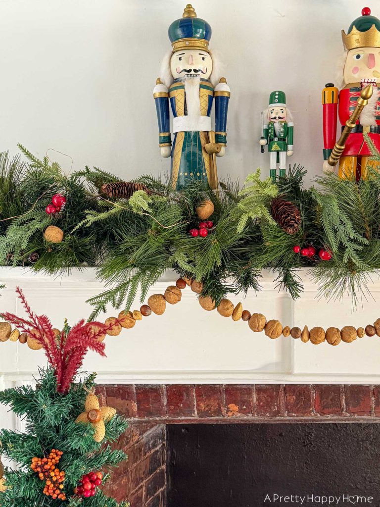 green holiday garland adorning a white fireplace mantel with brick surround. There is a hand strung, in-shell nut garland hanging below the green garland. On top of the mantel are colorful nutcrackers. All a part of the blog post five ways to decorate with nuts for christmas.