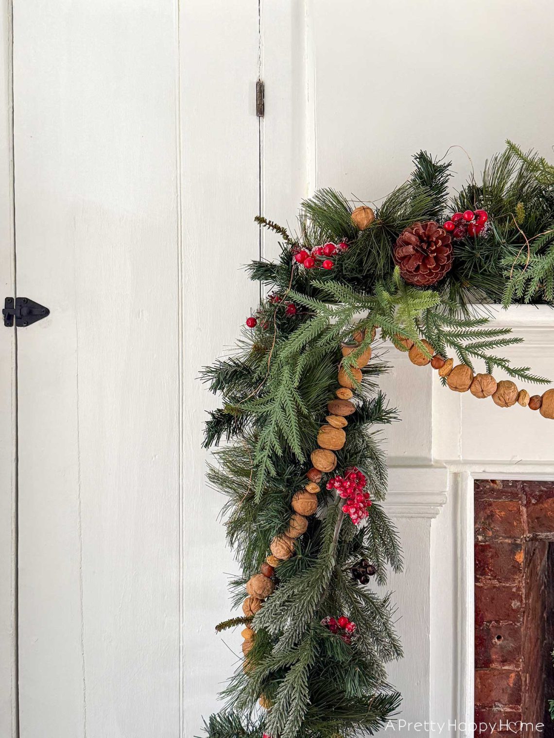 christmas garland with greenery and it is wrapped with a handmade nut garland of in-shell walnuts, almonds, and hazelnuts. the garland is draped over a white fireplace with brick surround and is featured on the blog post five ways to decorate with nuts for christmas
