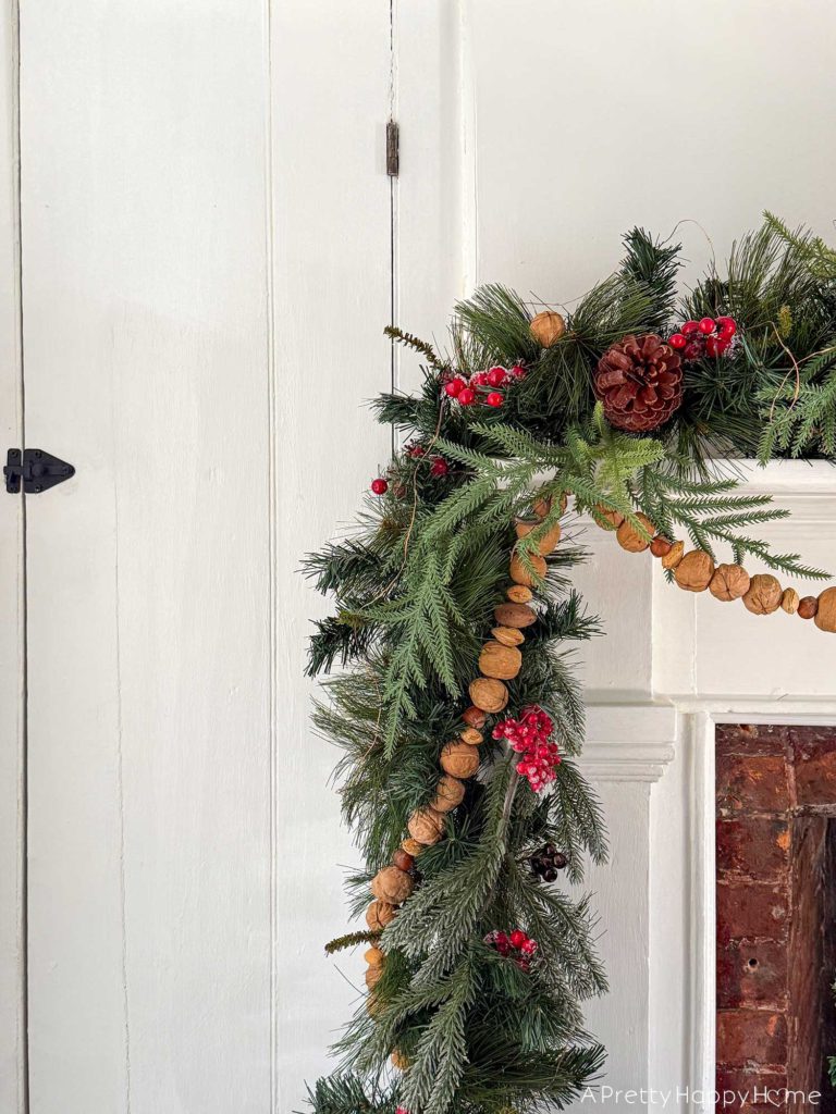 christmas garland with greenery and it is wrapped with a handmade nut garland of in-shell walnuts, almonds, and hazelnuts. the garland is draped over a white fireplace with brick surround and is featured on the blog post five ways to decorate with nuts for christmas