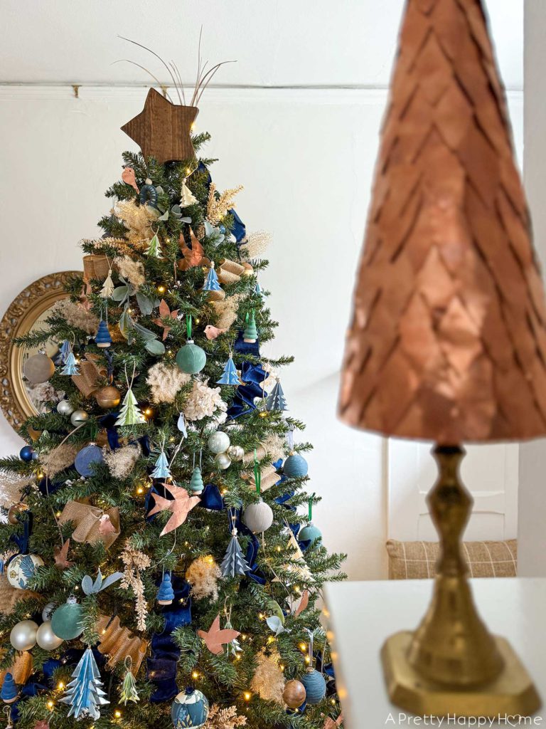 blue and metallic christmas tree decorated with small blue paper tree ornaments, faux velvet baubles in blue and green, small faux velvet tree ornaments in blue and green, pampas grass, blue and tan ribbon, kimekori ornaments, and a wood star topper. The tree is in front of an ornate oval mirror. There is a mini copper christmas tree sitting in the foreground on the living room mantel.