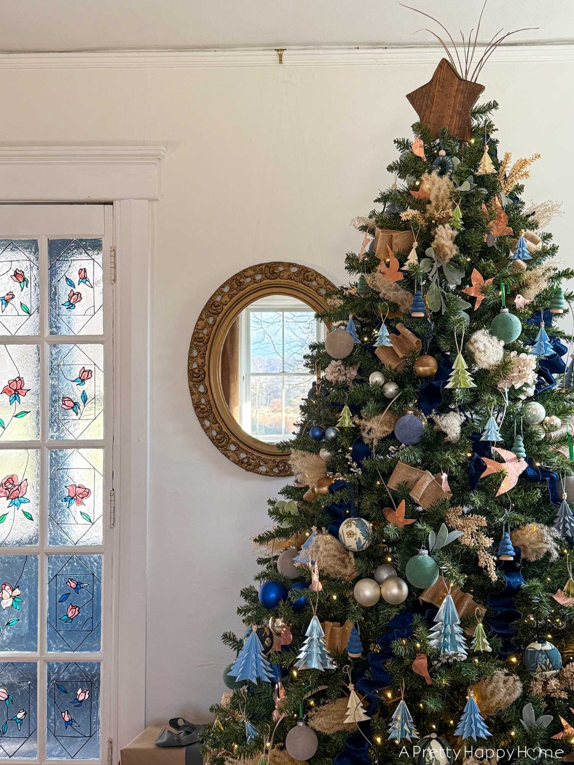 blue and metallic christmas tree decorated with small blue paper tree ornaments, faux velvet baubles in blue and green, small faux velvet tree ornaments in blue and green, pampas grass, blue and tan ribbon, kimekori ornaments, and a wood star topper. The tree is in front of an ornate oval mirror and stained glass french doors.