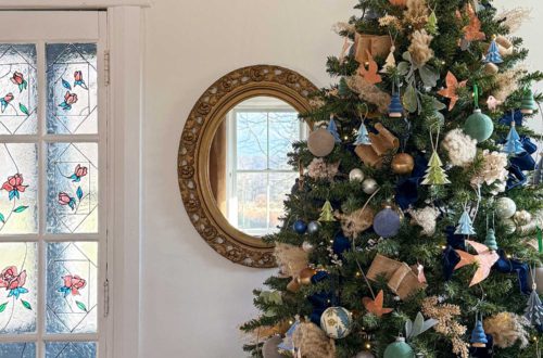 blue and metallic christmas tree decorated with small blue paper tree ornaments, faux velvet baubles in blue and green, small faux velvet tree ornaments in blue and green, pampas grass, blue and tan ribbon, kimekori ornaments, and a wood star topper. The tree is in front of an ornate oval mirror and stained glass french doors.