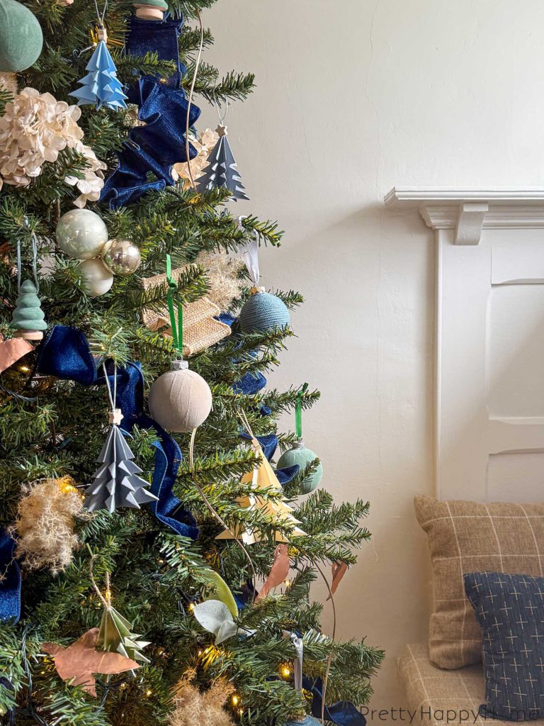 blue and metallic christmas tree decorated with small blue paper tree ornaments, faux velvet baubles in blue and green, small faux velvet tree ornaments in blue and green, pampas grass, blue and tan ribbon, kimekori ornaments, and a wood star topper. The tree is next to a built in window bench seat.