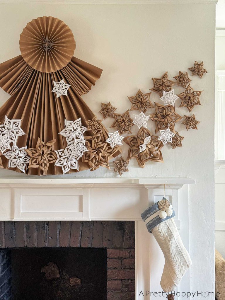 white fireplace with black brick surround decorated for christmas with a 4 foot tall folded giant paper angel made of brown kraft paper. Along the bottom of the angel and fanning out on the sides of her dress are fifty paper snowflakes made from white paper and brown kraft paper. Hanging under the mantel are two white and blue christmas stockings.