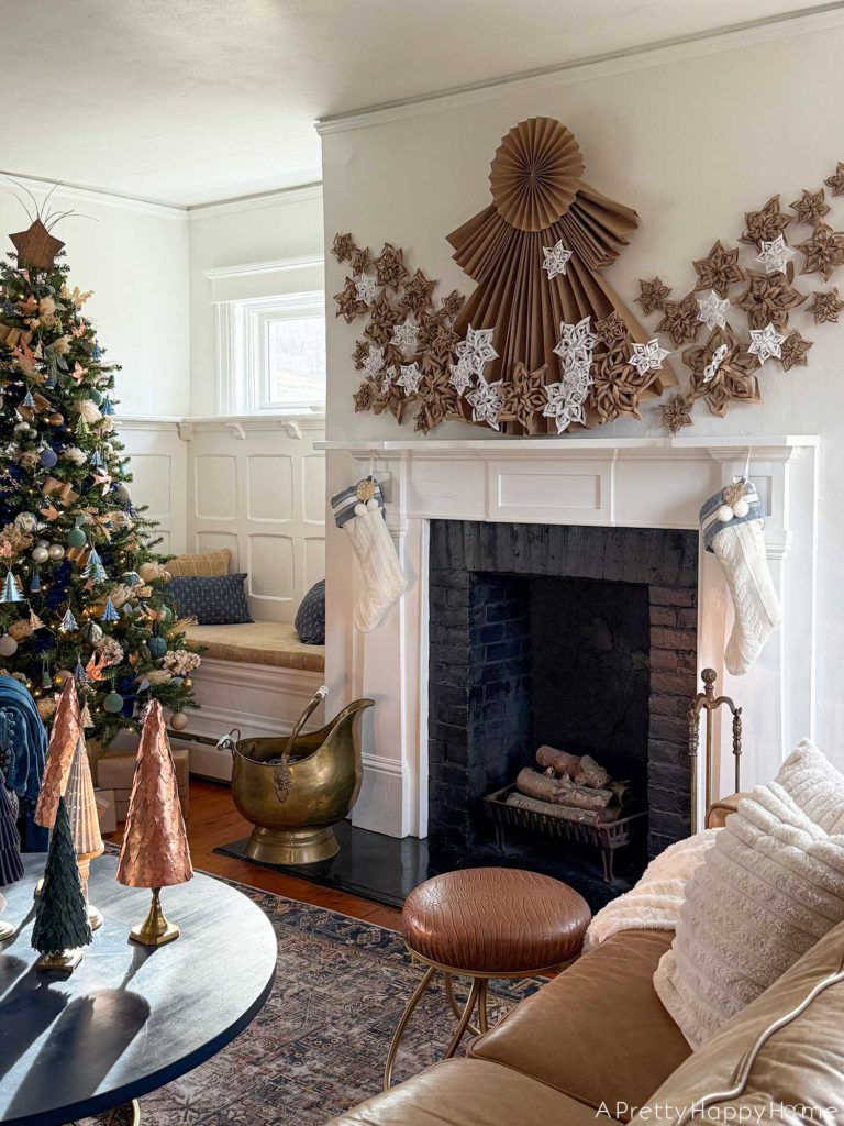 white fireplace with black brick surround decorated for christmas with a 4 foot tall folded giant paper angel made of brown kraft paper. Along the bottom of the angel and fanning out on the sides of her dress are fifty paper snowflakes made from white paper and brown kraft paper. Hanging under the mantel are two white and blue christmas stockings. There's a brass ash bucket in front of the fireplace and a brass fire tool set on the right hand side in front of the fireplace. There's a Christmas tree decorated in blue and green ornaments to the left of the fireplace. In front of the fireplace is a round blue coffee table with six mini Christmas trees sitting on it. Two of the tree are copper, one is white ceramic, one is blue flocked, two are made from green leather.
