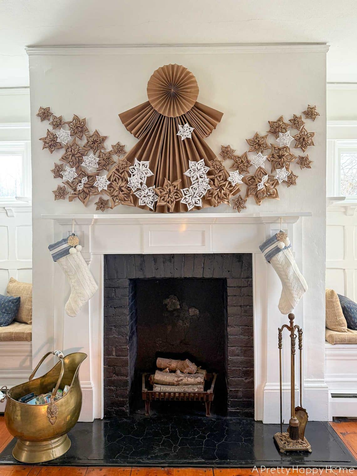 white fireplace with black brick surround decorated for christmas with a 4 foot tall folded giant paper angel made of brown kraft paper. Along the bottom of the angel and fanning out on the sides of her dress are fifty paper snowflakes made from white paper and brown kraft paper. Hanging under the mantel are two white and blue christmas stockings. There's a brass ash bucket in front of the fireplace and a brass fire tool set on the right hand side in front of the fireplace.