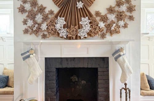 white fireplace with black brick surround decorated for christmas with a 4 foot tall folded giant paper angel made of brown kraft paper. Along the bottom of the angel and fanning out on the sides of her dress are fifty paper snowflakes made from white paper and brown kraft paper. Hanging under the mantel are two white and blue christmas stockings. There's a brass ash bucket in front of the fireplace and a brass fire tool set on the right hand side in front of the fireplace.
