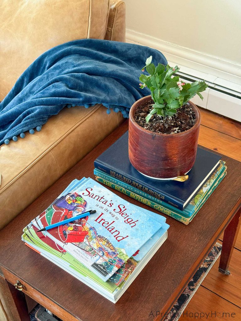 christmas around our home 2025 showing a candid snapshot of a side table next to a butter-colored leather couch with a blue throw blanket draped over the arm of the couch. On the table are two stacks of books. The first stack has a christmas cactus in a wood pot. The cactus is starting to bloom. The second stack of books is a collection of Santa's Sleigh books. The title of the book sitting on top of the pile is Santa's sleigh is coming to ireland. There's a blue mechanical pencil sitting on top of the stack of books, likely left there after a kid did homework.