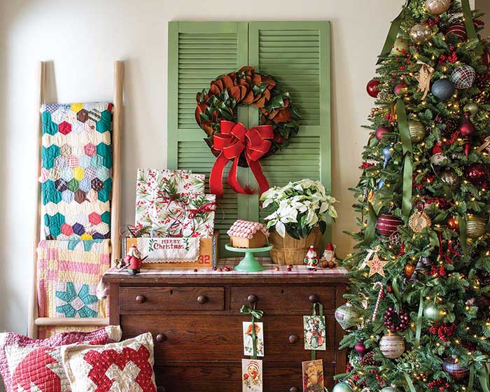 vignette of a farmhouse Christmas. there's a christmas tree on the right decorated with colorful ornaments. There's a wood dresser with green shutters leaning above it. On the shutters are a green wreath with red bow. There's a blanket ladder to the left of the dresser with colorful quilts, and a container of colorful quilted pillows below that. Photography by John O'Hagan, Styling by Maghan Armstrong via The Cottage Journal on the happy list