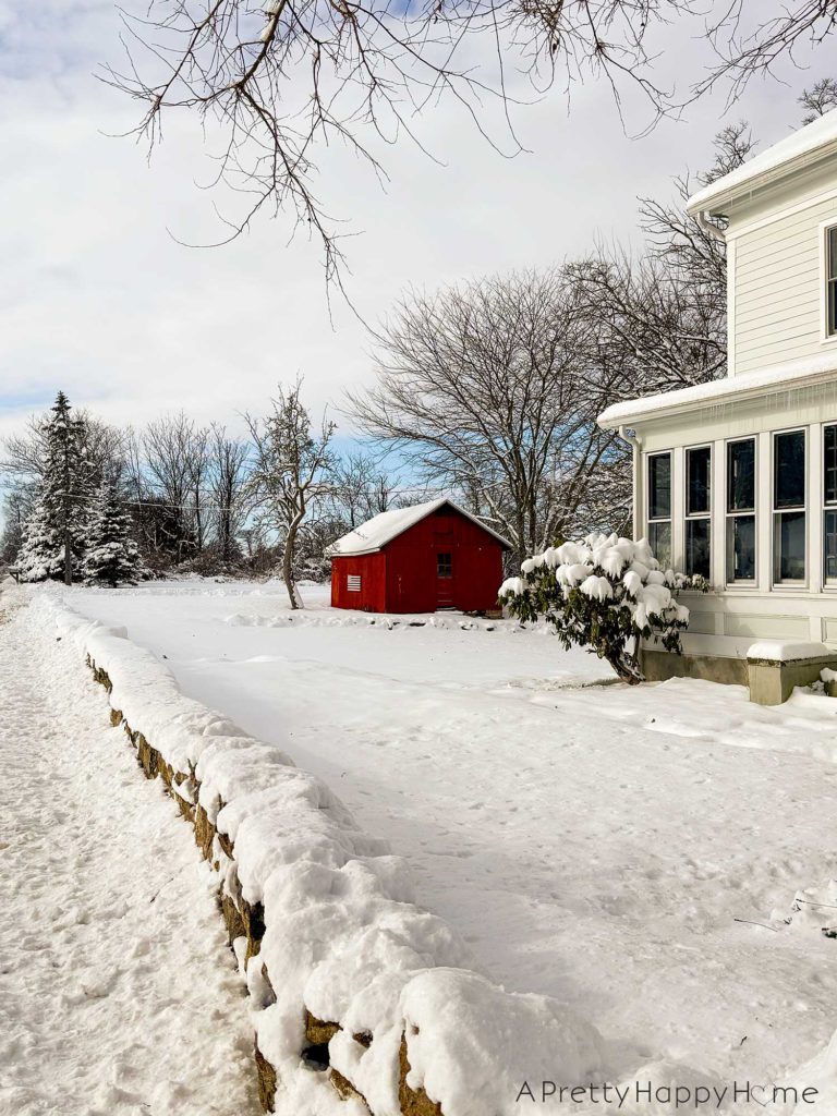 White farmhouse covered in snow. A snow covered low rock wall is in front of the house. There's a small red bard in the distance.