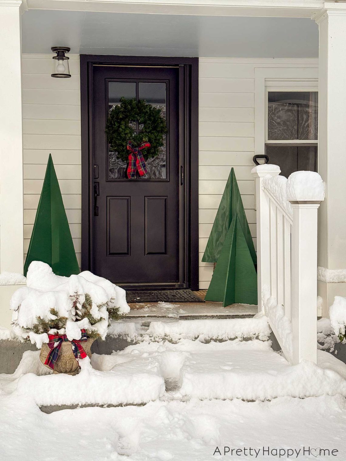 Front porch of white house with black front door. There is a green wreath with a plaid bow hanging on the front door. There are four trees of varying sized made out of plywood and painted green on either side of the door. The steps leading up to the porch are covered in snow.