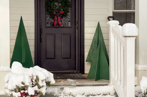 Front porch of white house with black front door. There is a green wreath with a plaid bow hanging on the front door. There are four trees of varying sized made out of plywood and painted green on either side of the door. The steps leading up to the porch are covered in snow.