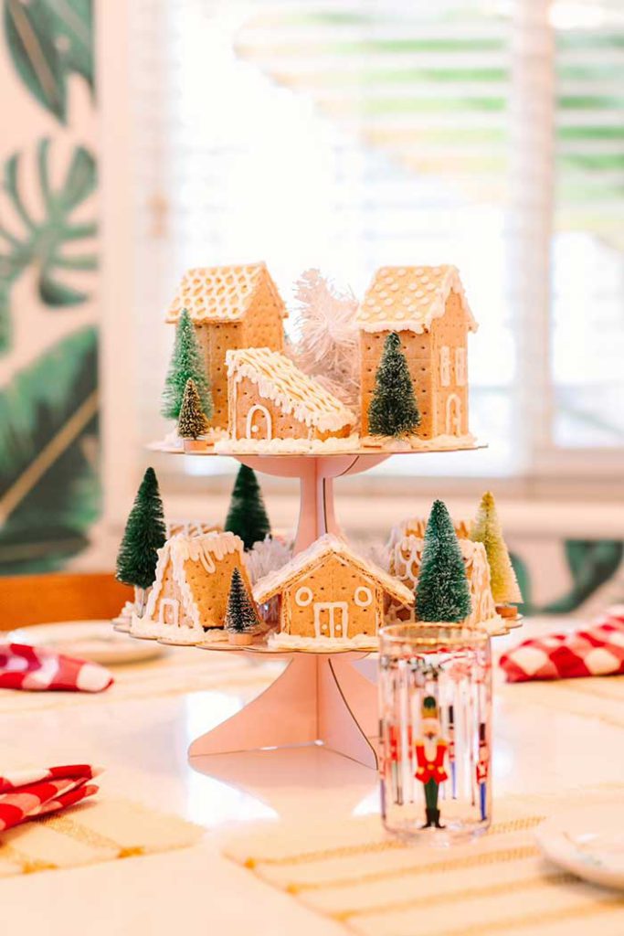 A Christmas centerpiece of a graham cracker village on a two-tiered cake stand. The houses are decorated with white icing to look like mini gingerbread houses. There are green bottle brush trees interspersed with the houses. Image from Lovely Indeed and shown here on the Happy List.