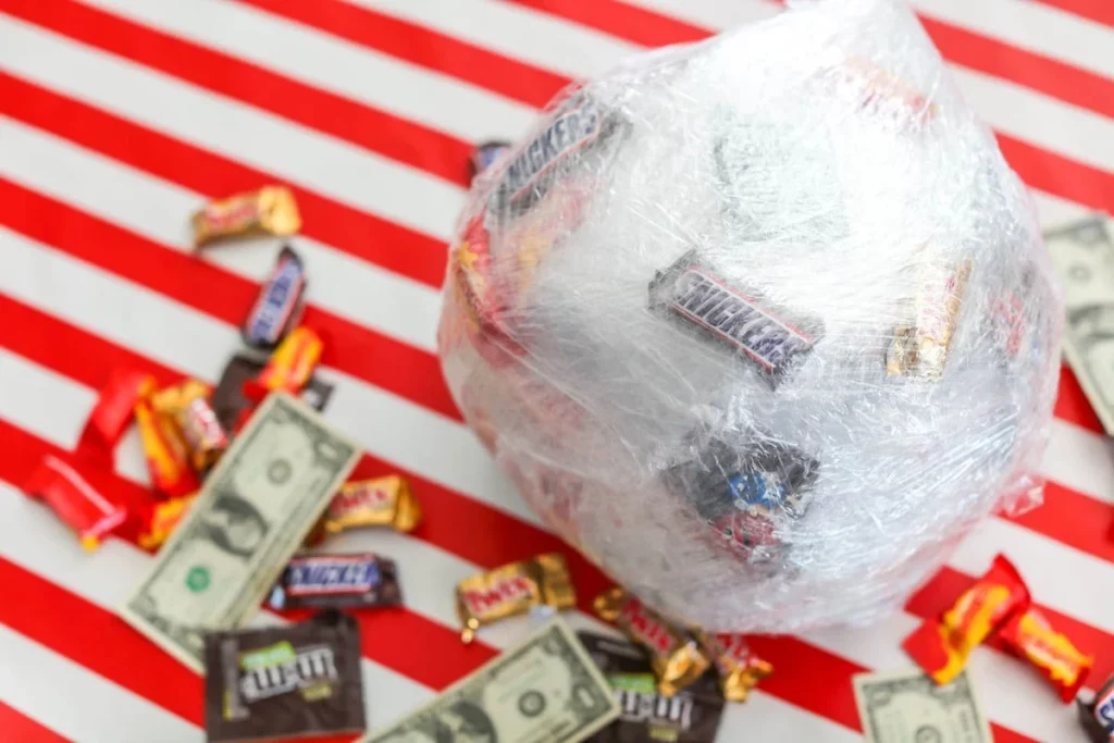 saran wrap ball game. image shows a large ball of saran wrap holding small prizes in place like candy and gift cards. The ball is sitting on red and white striped background and it surrounded by potential game prizes like cash and candy. Image by Lil' Luna and shown here on the happy list