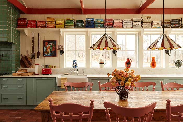 photo of cookbook author dan pelosi's colorful kitchen. there are mint green cabinets, cookbooks stacked in rainbow order on a shelf above a large kitchen window, a wood table with bright tiffany glass pendants hanging over it. Image via Domino shown here on the happy list. photographer is Kate S Jordan and styling by Kate Berry.