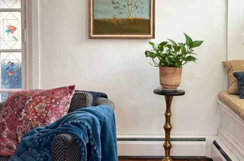living room vignette focusing on a vintage paragon needlecraft called birch in the wind hanging on the wall. There's a low blue chair sitting in front of the needlepoint. There's also a pothos plant sitting on a wood and brass plant stand in front of the needlepoint, which was thrifted fiber art.