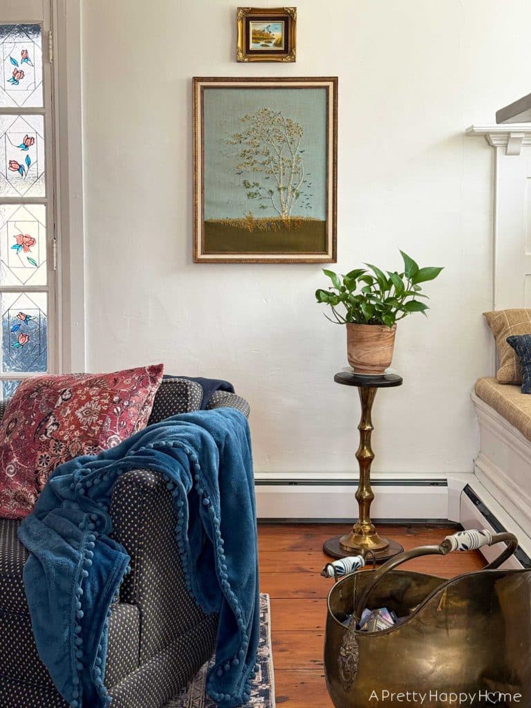 living room vignette focusing on a vintage paragon needlecraft called birch in the wind hanging on the wall. There's a low blue chair sitting in front of the needlepoint. There's also a pothos plant sitting on a wood and brass plant stand in front of the needlepoint, which was thrifted fiber art.