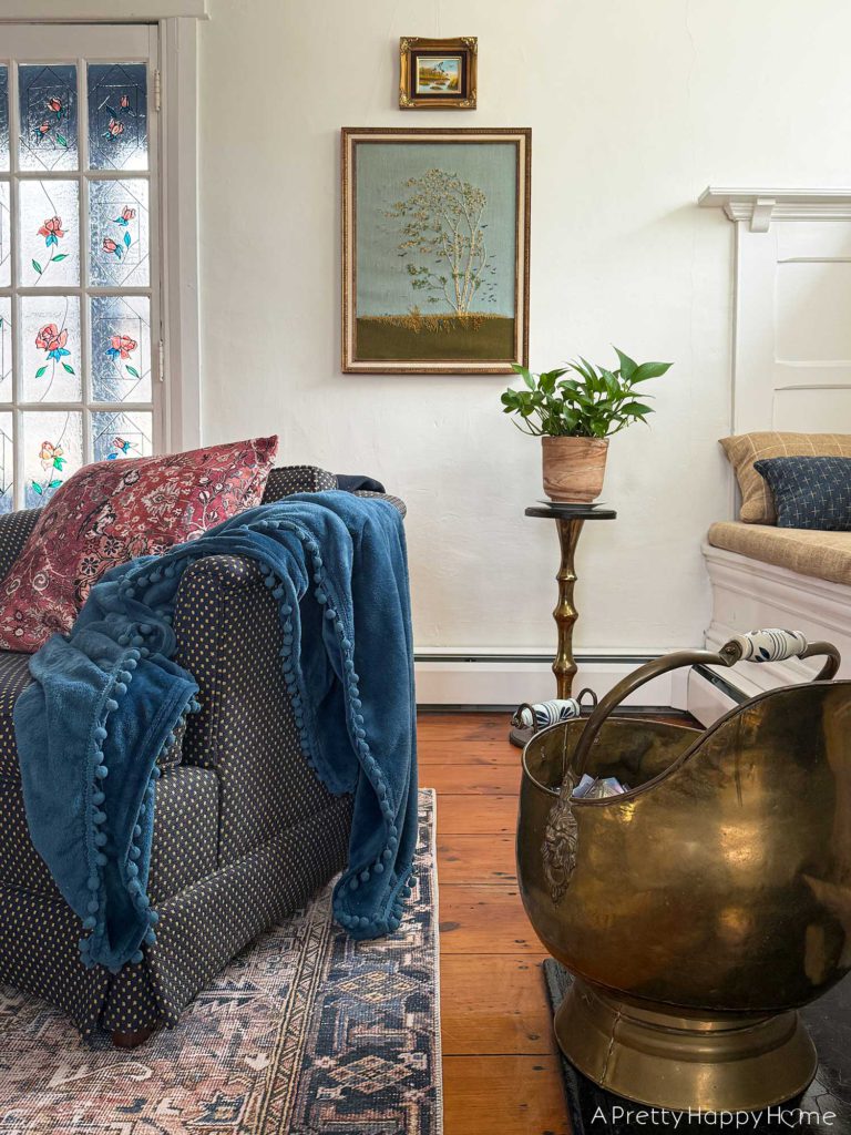 living room vignette focusing on a vintage paragon needlecraft called birch in the wind hanging on the wall. There's a low blue chair sitting in front of the needlepoint. There's also a pothos plant sitting on a wood and brass plant stand in front of the needlepoint, which was thrifted fiber art.