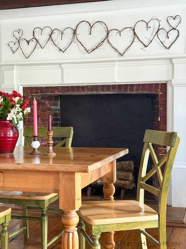 image showing a white fireplace with a grapevine heart garland hanging above the fireplace. The grapevines are a variety of sizes. In the foreground is a wood dining room table with a red vase holding red and white alstroemerias. There are also two brass candle holders on the table with red and pink candles. The chairs around the dining room table are green and natural wood. The picture is showcasing how to DIY a grapevine heart garland.