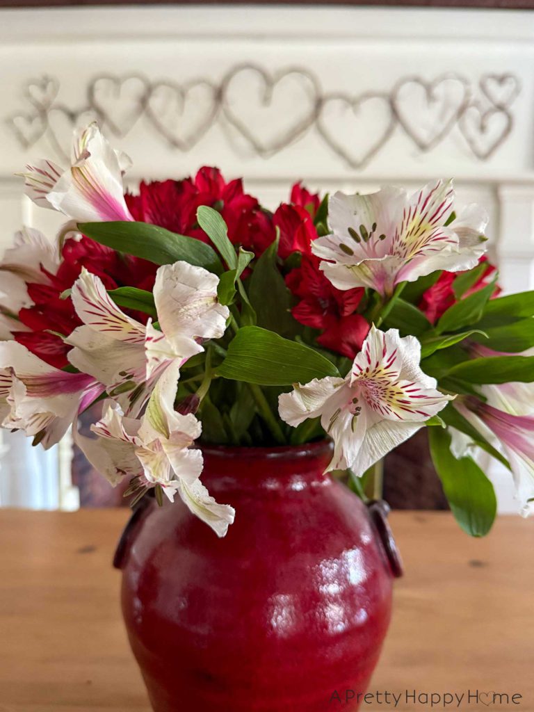 red pottery vase holding red and white flowers sitting on a table with a grapevine heart garland in the background