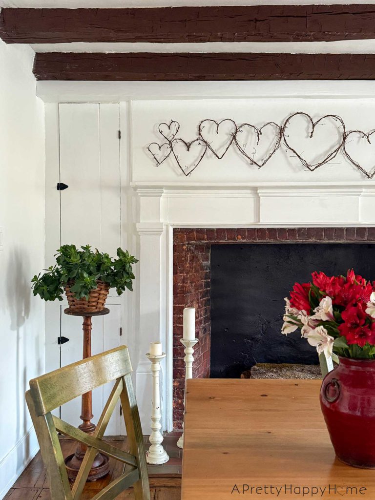 image showing a white fireplace with a grapevine heart garland hanging above the fireplace. The grapevines are a variety of sizes. In the foreground is a wood dining room table with a red vase holding red and white alstroemerias. The picture is showcasing how to DIY a grapevine heart garland.