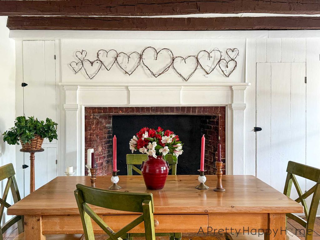 image showing a white fireplace with a grapevine heart garland hanging above the fireplace. The grapevines are a variety of sizes. In the foreground is a wood dining room table with a red vase holding red and white alstroemerias. There are also four brass candle holders on the table with red and pink candles. The picture is showcasing how to DIY a grapevine heart garland.