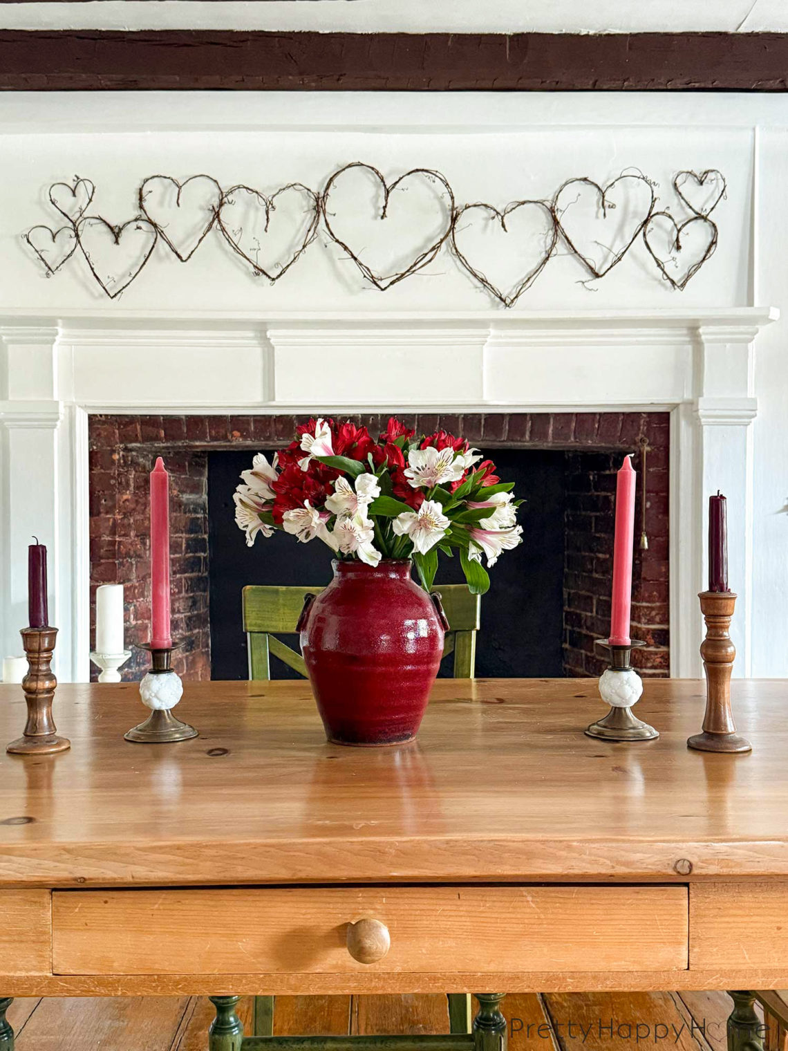 image showing a white fireplace with a grapevine heart garland hanging above the fireplace. The grapevines are a variety of sizes. In the foreground is a wood dining room table with a red vase holding red and white alstroemerias. There are also four brass candle holders on the table with red and pink candles. The picture is showcasing how to DIY a grapevine heart garland.