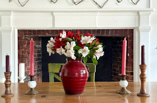 image showing a white fireplace with a grapevine heart garland hanging above the fireplace. The grapevines are a variety of sizes. In the foreground is a wood dining room table with a red vase holding red and white alstroemerias. There are also four brass candle holders on the table with red and pink candles. The picture is showcasing how to DIY a grapevine heart garland.