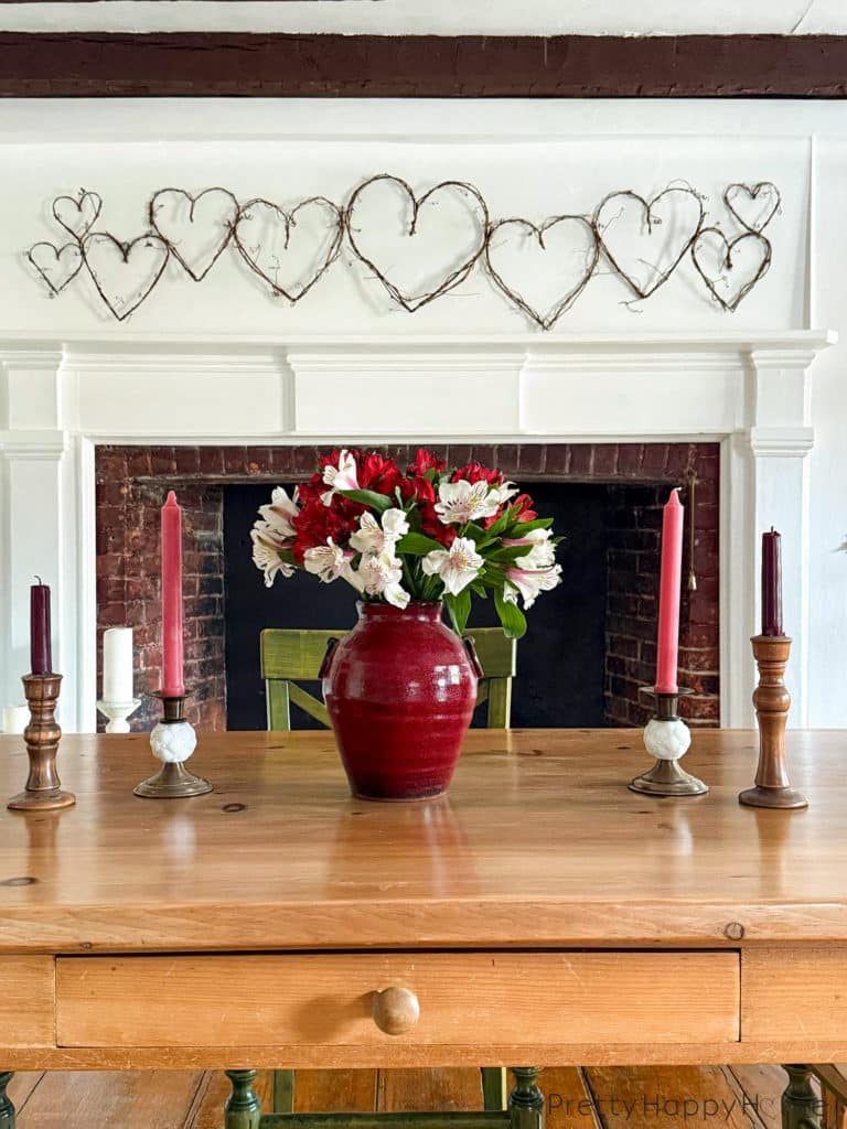 image showing a white fireplace with a grapevine heart garland hanging above the fireplace. The grapevines are a variety of sizes. In the foreground is a wood dining room table with a red vase holding red and white alstroemerias. There are also four brass candle holders on the table with red and pink candles. The picture is showcasing how to DIY a grapevine heart garland.