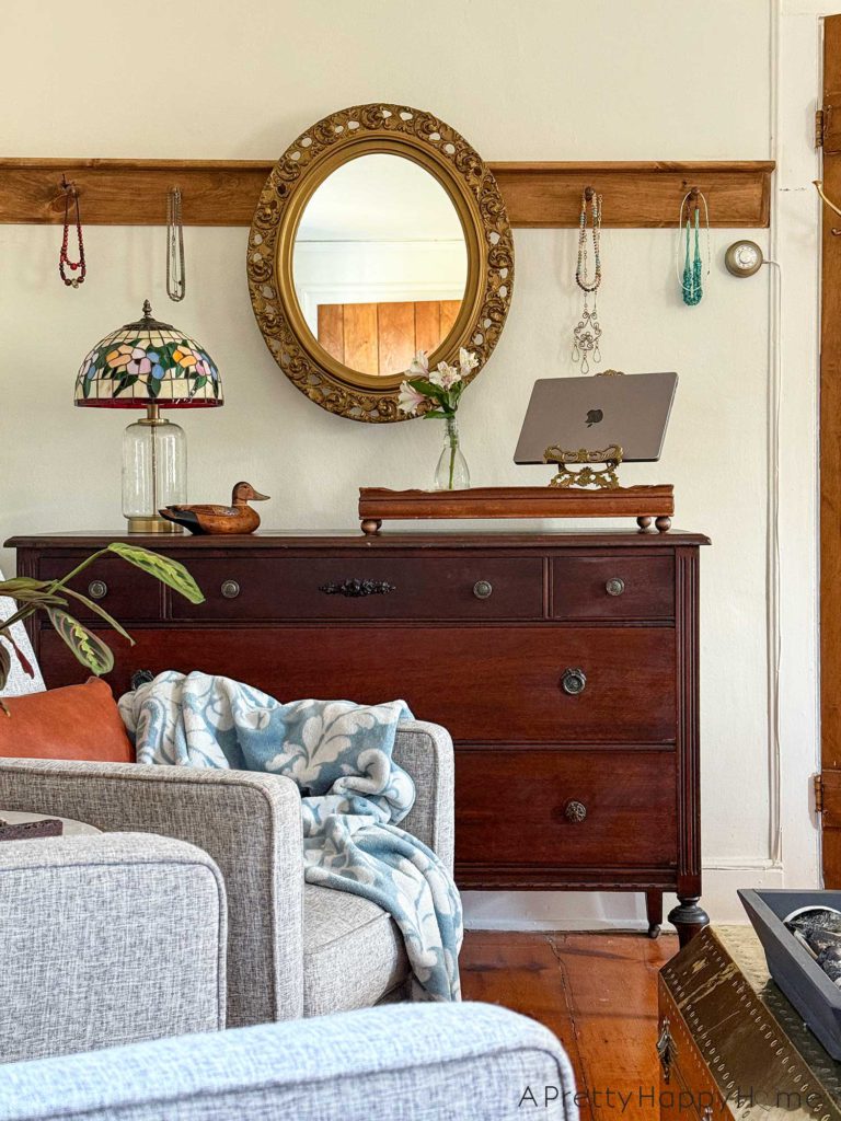 ornate oval mirror hanging from a wood peg rail. There are necklaces on the pegs on either side of the mirror. The mirror hangs above a wood dresser. On the dresser is a tiffany glass lamp, a wood duck, a wood tray with a bouquet of flowers and a laptop stand. In front of the dresser is a seating area with landscape paintings on the wall, grey recliner chairs, a prayer plant, and a light blue throw. The image is showcasing how to hang a mirror from a peg rail.