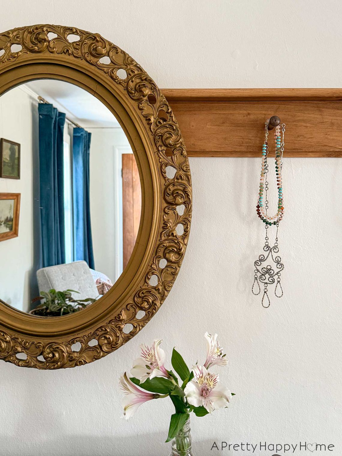 Ornate oval mirror hanging from a wood peg rail. One peg is showing a silver necklace and a colorful beaded necklace hanging from it. There is a clear vase of white Alstroemeria below the picture. A window with navy blue curtains is reflected in the mirror. So, yes, you can hang a mirror from a peg rail.