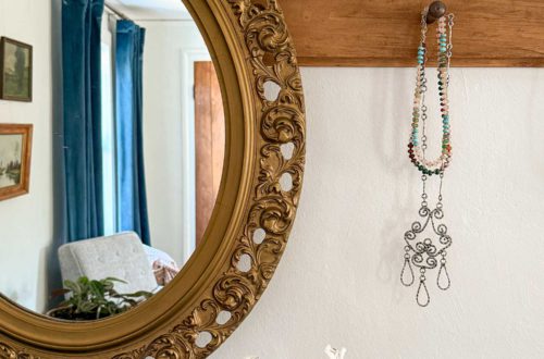 Ornate oval mirror hanging from a wood peg rail. One peg is showing a silver necklace and a colorful beaded necklace hanging from it. There is a clear vase of white Alstroemeria below the picture. A window with navy blue curtains is reflected in the mirror. So, yes, you can hang a mirror from a peg rail.