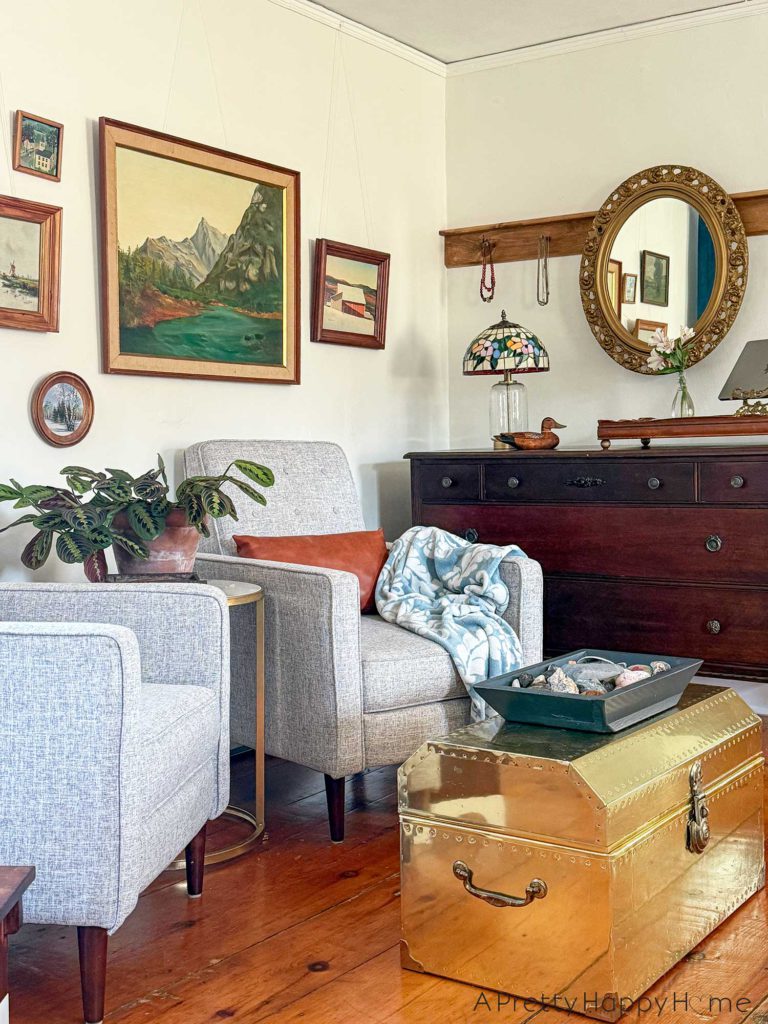 ornate oval mirror hanging from a wood peg rail. There are necklaces on the pegs on either side of the mirror. The mirror hangs above a wood dresser. On the dresser is a tiffany glass lamp, a wood duck, a wood tray with a bouquet of flowers and a laptop stand. In front of the dresser is a seating area with landscape paintings on the wall, grey recliner chairs, a prayer plant, and a light blue throw. The image is showcasing how to hang a mirror from a peg rail.