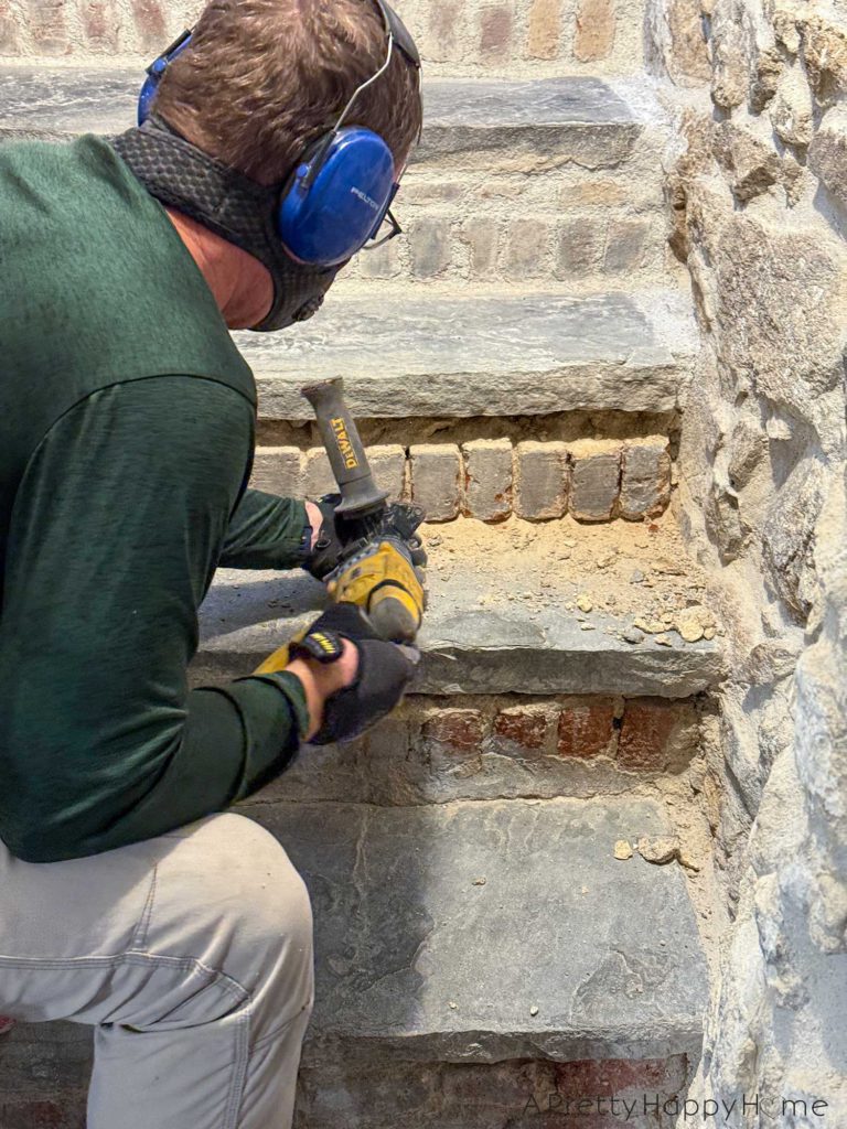 man wearing safety gear chipping out old lime mortar from around bricks on stair risers to a basement. He's using a chisel attachment on a hammer drill to remove the old mortar.